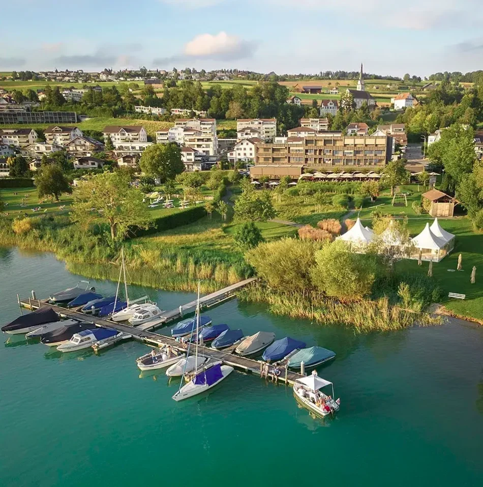 Sempachersee bei Eich aus der Luft: Bootssteg mit Segel- und Motorbooten, dahinter Seegarten mit Zeltpavillons und Hotel